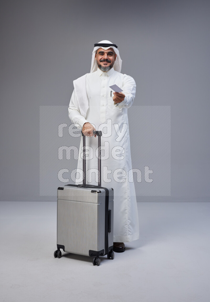 Saudi man wearing Thob and white Shomag standing holding Travel bag and ATM card on Gray background