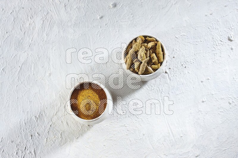 وعاءان فخاريان بيج ممتلئان ببودرة الكركم وأصابع الكركم المجففة على خلفية بيضاء