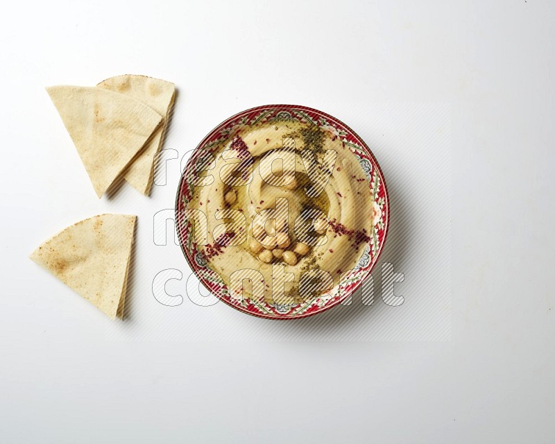 Hummus in a red plate with patterns garnished with zaatar & sumak on a white background