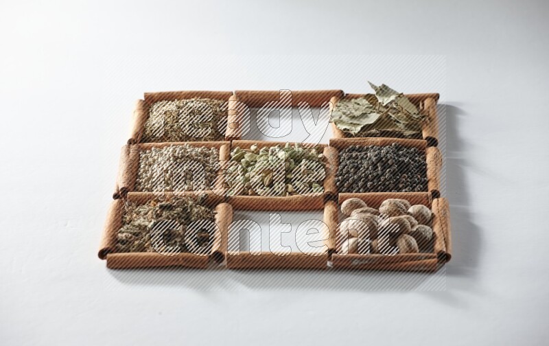 9 مربعات من أعواد القرفة ممتلئة بالهيل في المنتصف محاطة بجوزة الطيب والملح والريحان المجفف والفلفل الأبيض والكمون وبهار الفلفل الحلو وأوراق الغار والفلفل الأسود على أرضية بيضاء