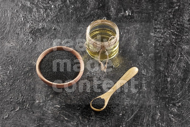 A wooden bowl and spoon full of black seeds and a glass jar of black seeds oil on a textured black flooring