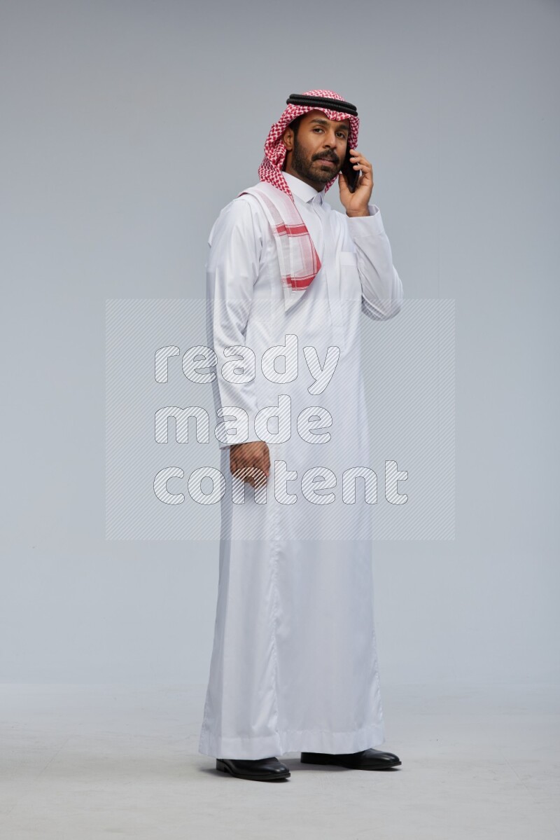 Saudi man Wearing Thob and shomag standing talking on phone on Gray background