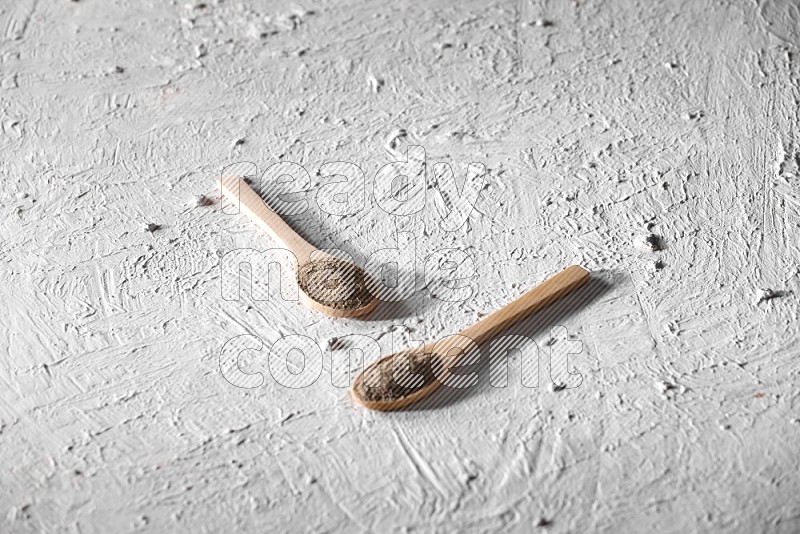 2 wooden spoons full of black pepper powder on a textured white background