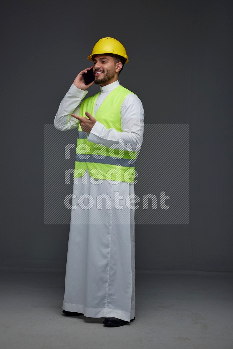 A Saudi man wearing Thobe with a yellow safety vest and white helmet standing and talking in the phone eye level on a gray background