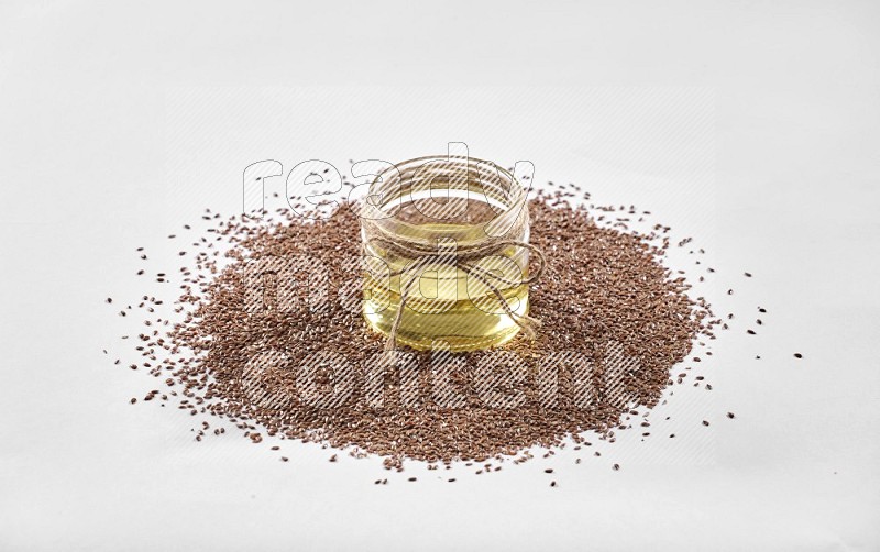 A glass jar full of flaxseeds oil surrounded by flax seeds on a white flooring