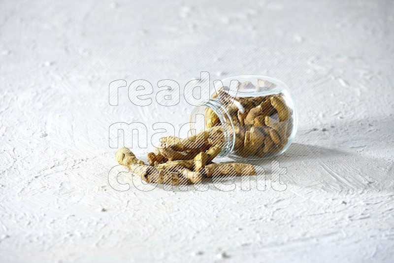 A flipped glass spice jar full of dried turmeric fingers and fell out it on textured white flooring