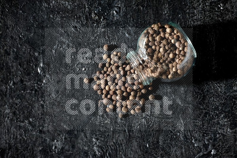 A flipped glass spice jar full of allspice whole balls and the balls spilled out of it on a textured black flooring