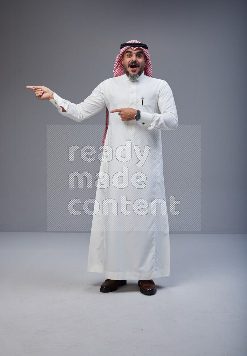 Saudi man Wearing Thob and red white Shomag standing interacting with the camera on Gray background