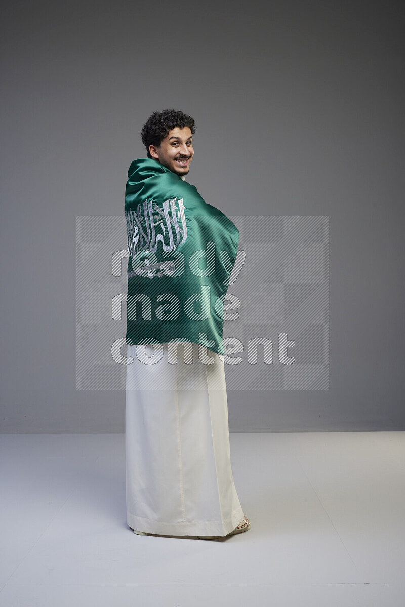 A Saudi man standing wearing thob wrapping big Saudi flag on gray background