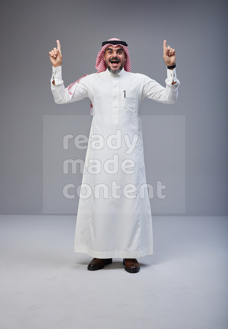 Saudi man Wearing Thob and red white Shomag standing interacting with the camera on Gray background