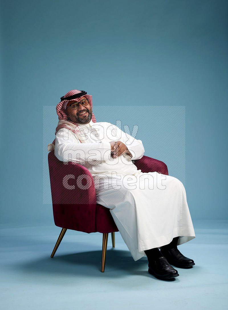 Saudi Man with shimag sitting on chair Interacting with the camera on blue background