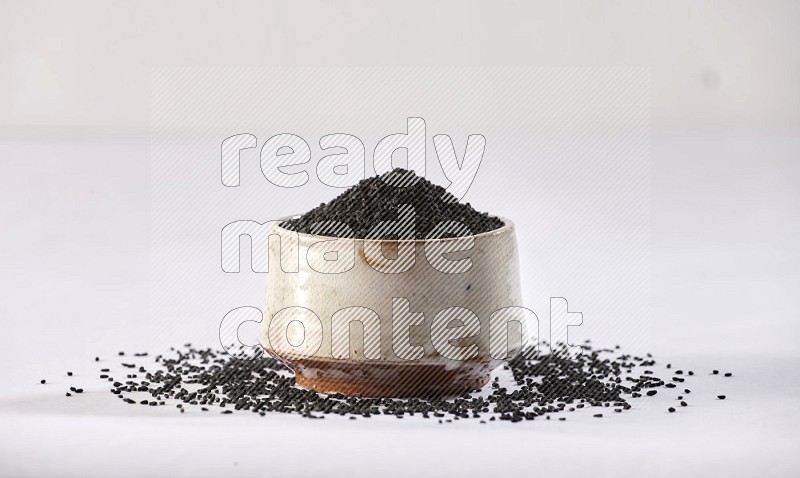 A beige pottery bowl full of black seeds and more seeds spread on a white flooring
