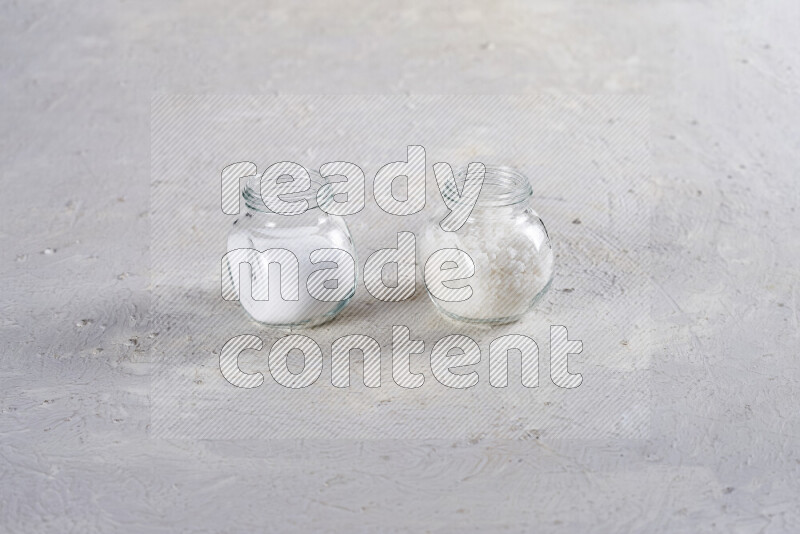 2 glass jars one is filled with table salt and the other with coarse sea salt on white background