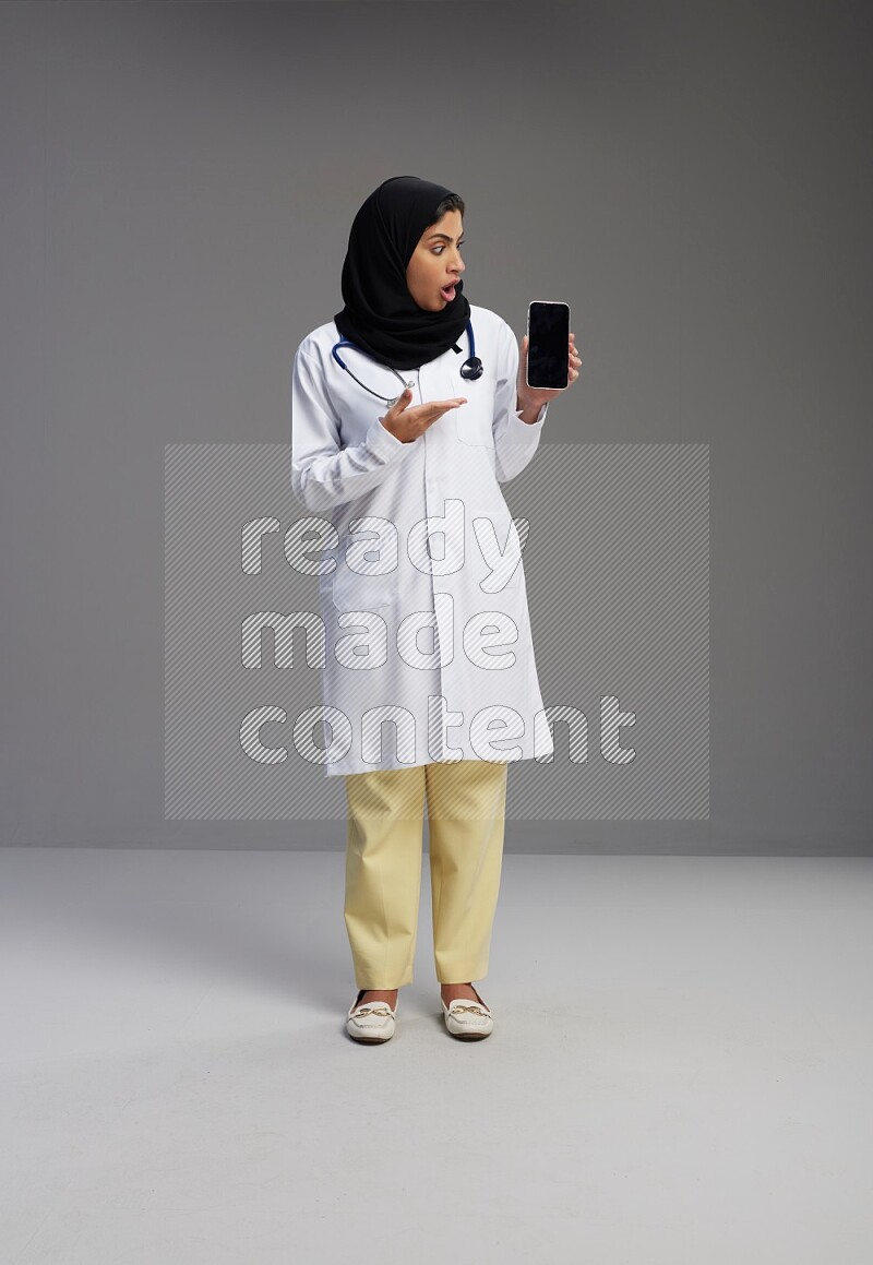 Saudi woman wearing lab coat with stethoscope standing showing phone to camera with sign in the back on Gray background