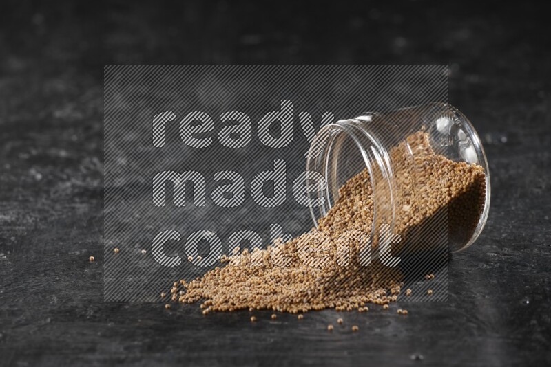 A glass jar full of mustard seeds and jar is flipped and seeds spread out on a textured black flooring