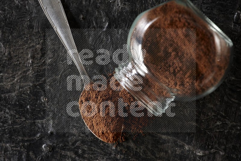 وعاء زجاجي للبهارات مقلوب وملعقة معدنية ممتلئان ببودرة القرنفل على أرضية سوداء