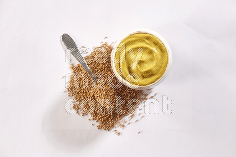 A beige pottery bowl full of mustard paste with mustard seeds underneath with a metal spoon on white flooring