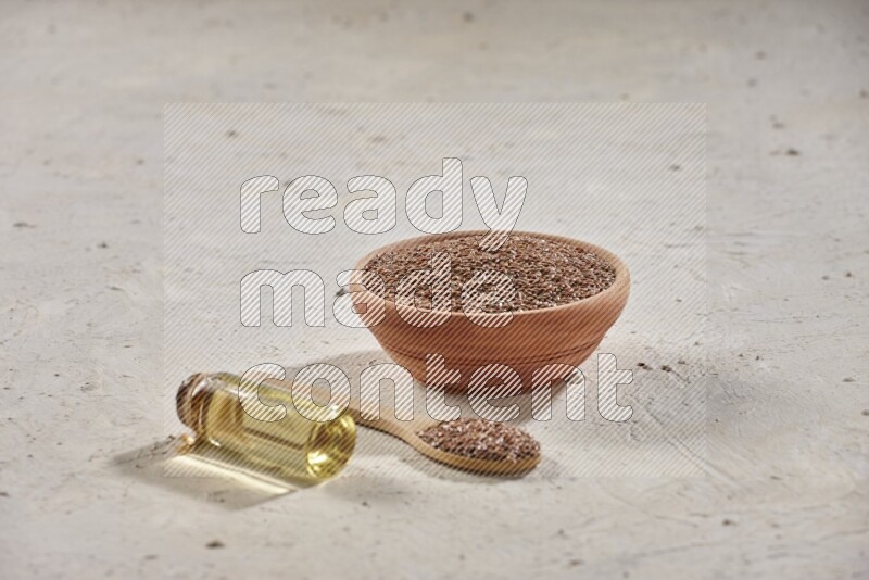 A wooden bowl and spoon full of flaxseeds with a bottle of flaxseeds oil on a textured white flooring