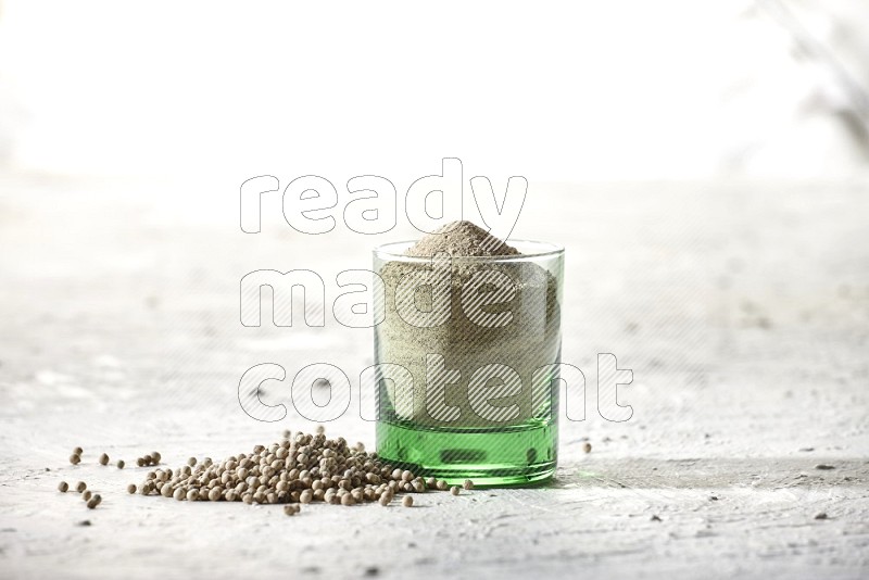 A green glass cup full of white pepper powder with white pepper beads on textured white flooring