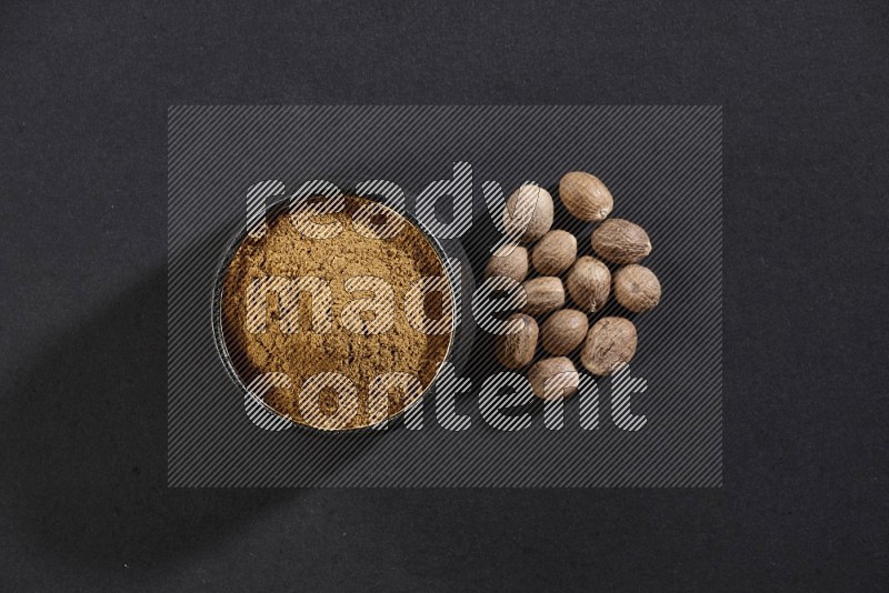 A black pottery bowl full of nutmeg powder with the seeds beside it on a black flooring in different angles