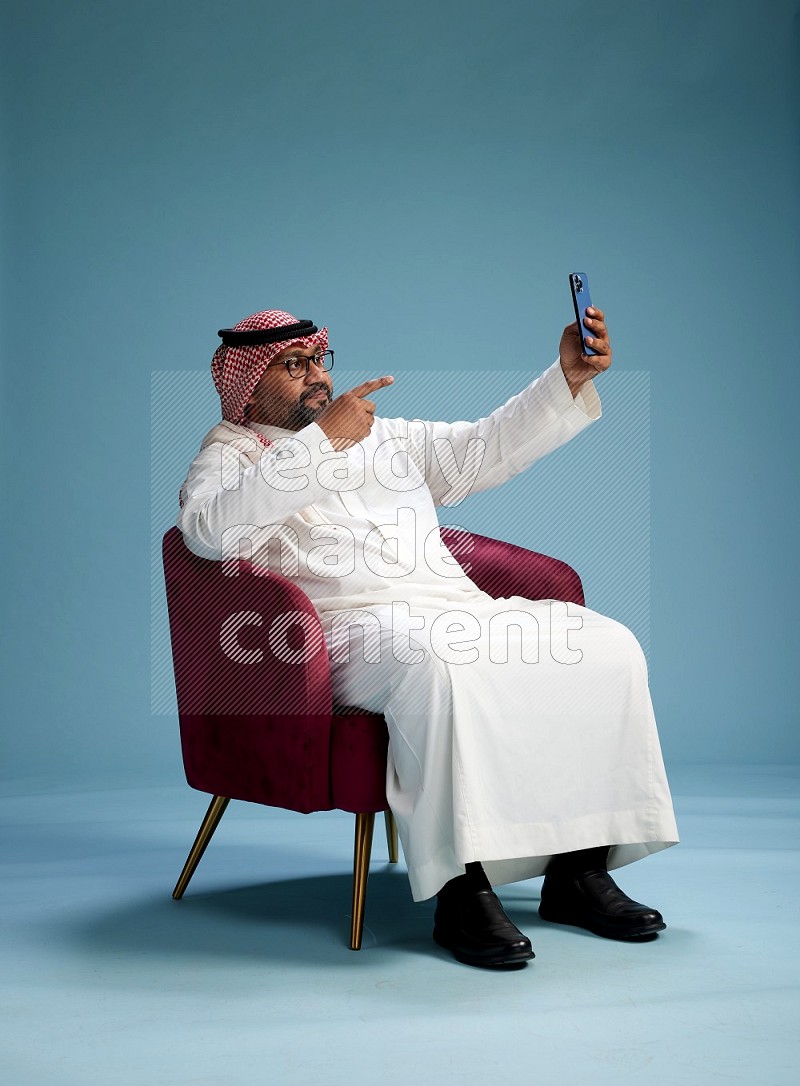 Saudi Man with shimag sitting on chair taking selfie on blue background