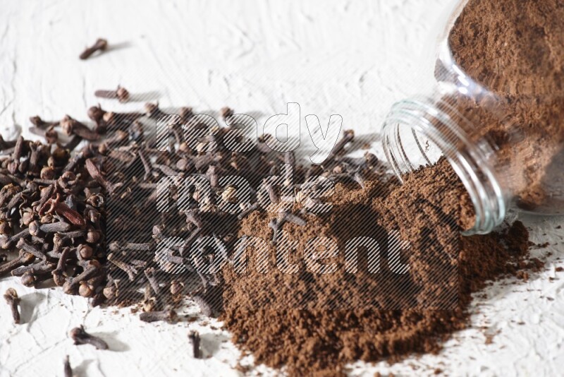 A glass spice jar full of cloves powder flipped and powder came out of it with cloves spread on textured white flooring