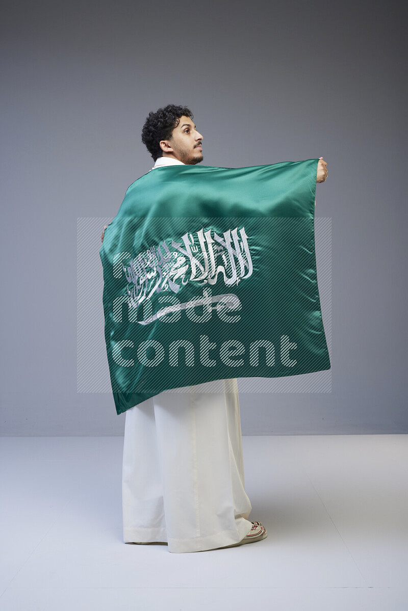 A Saudi man standing wearing thob holding big Saudi flag on gray background