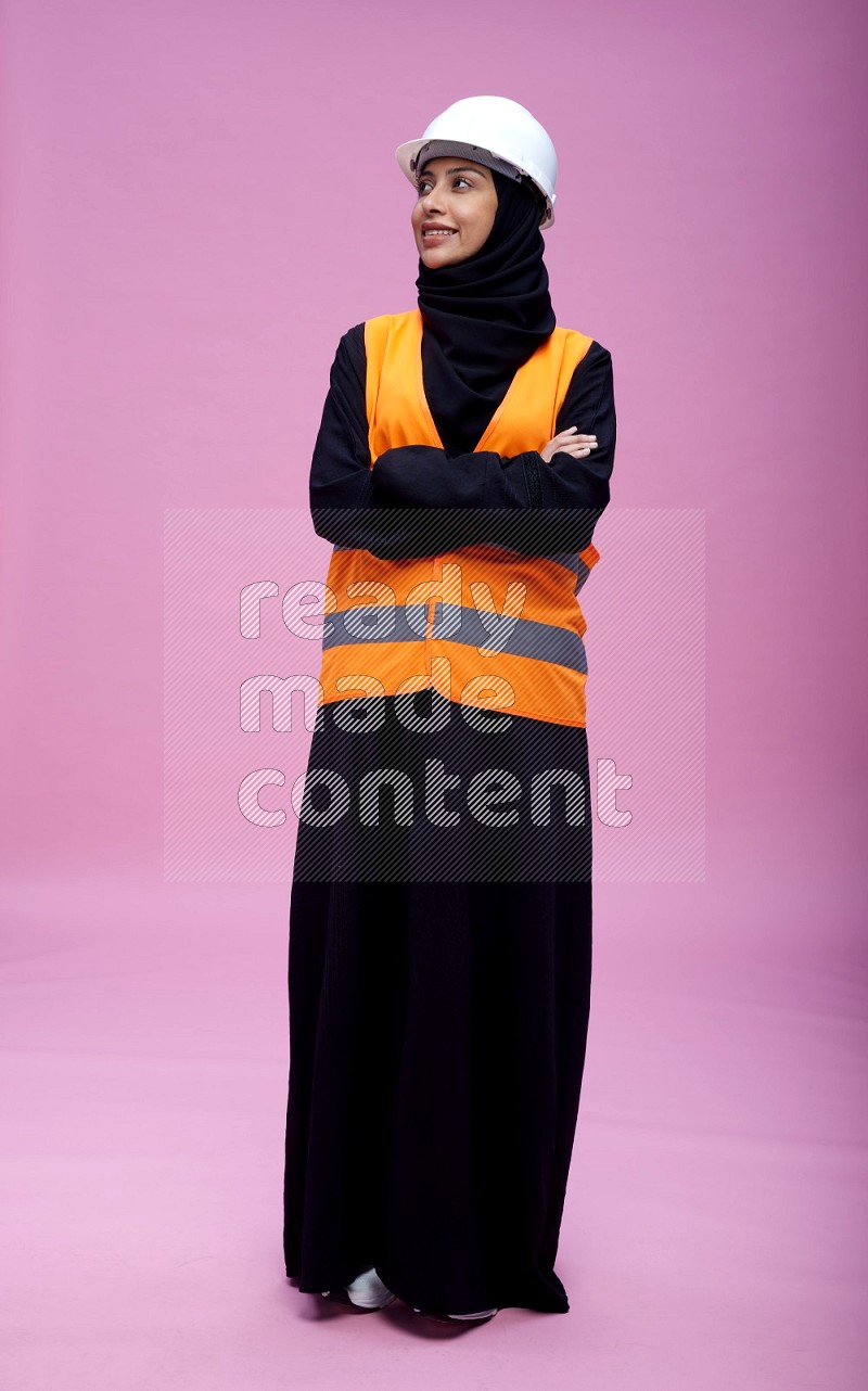 Saudi woman wearing Abaya with engineer vest and helmet standing with crossed arms on pink background