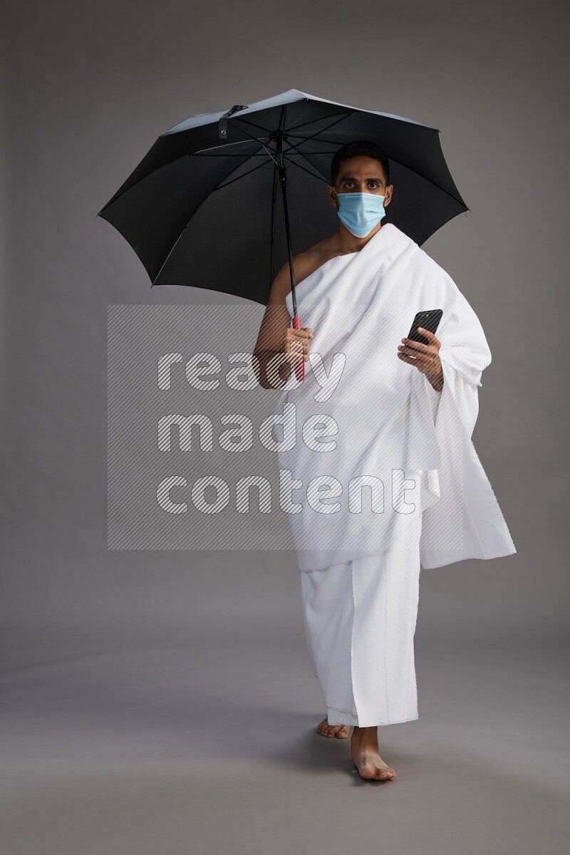 A man wearing Ehram and mask Standing holding umbrella on gray background on gray background