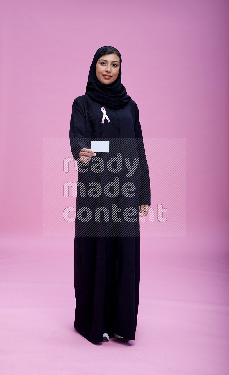 Saudi woman wearing pink ribbon on Abaya standing holding ATM card on pink background