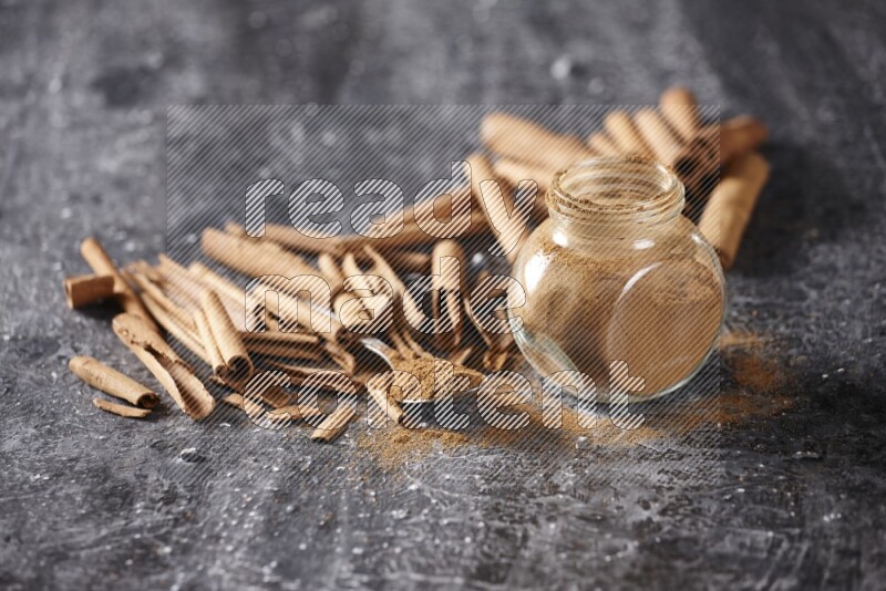 Herbal glass jar and a metal spoon full of cinnamon powder surrounded by cinnamon sticks on textured black background