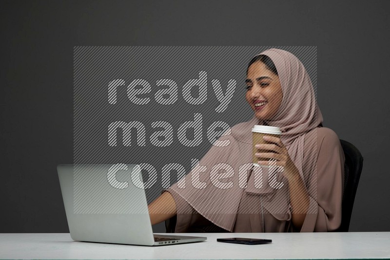 A Female Sitting on her desk drinking coffee on a Gray Background wearing Brown Abaya with Hijab
