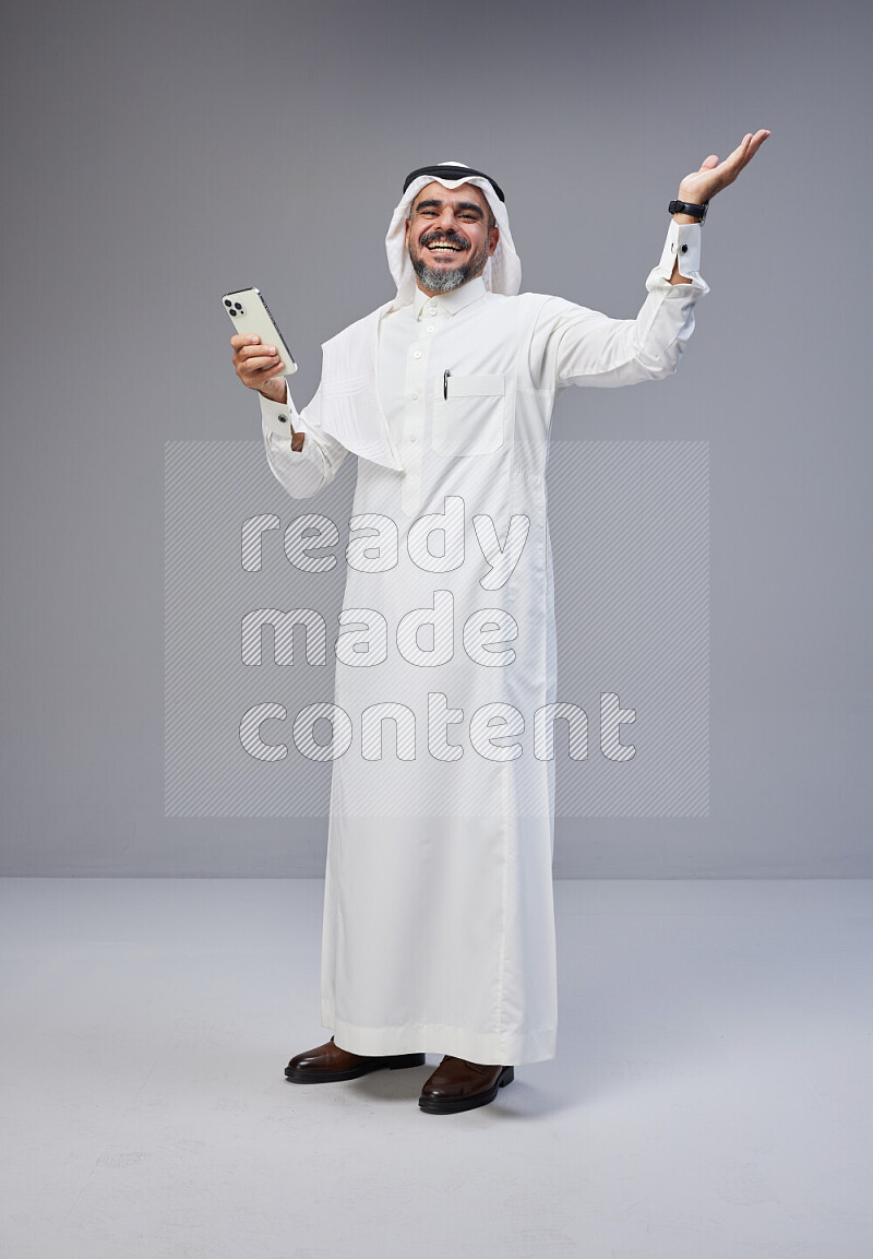 Saudi man Wearing Thob and white Shomag standing texting on phone on Gray background