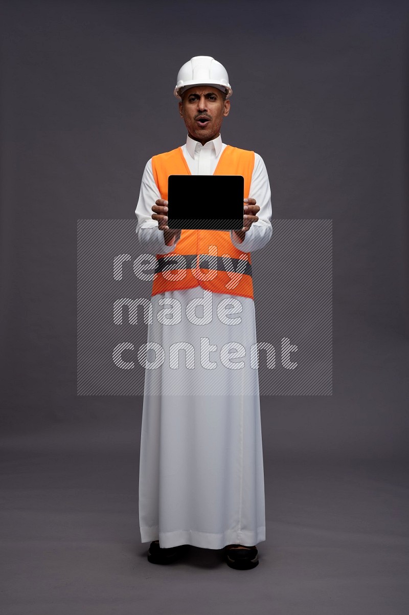 Saudi man wearing thob with engineer vest standing showing tablet to camera on gray background