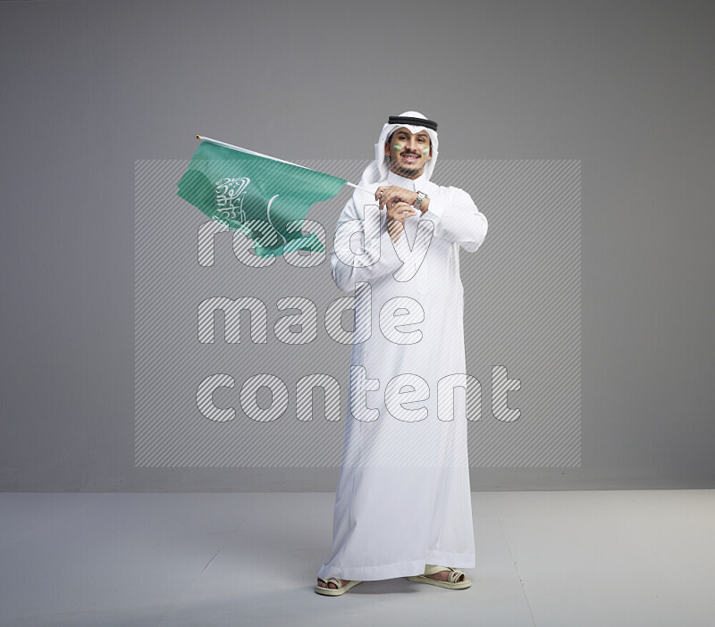 A Saudi man standing wearing thob and white shomag with face painting raising big Saudi flag on gray background