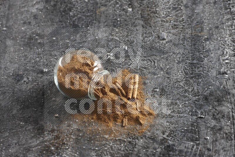 Flipped glass herbs jar full of cinnamon powder with cracked cinnamon sticks on a textured black background