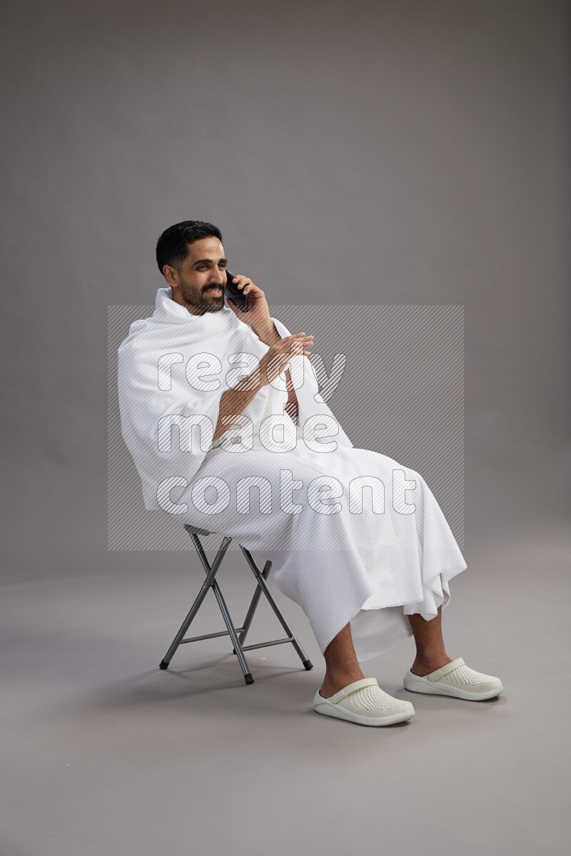 A man wearing Ehram sitting on chair talking on the phone on gray background