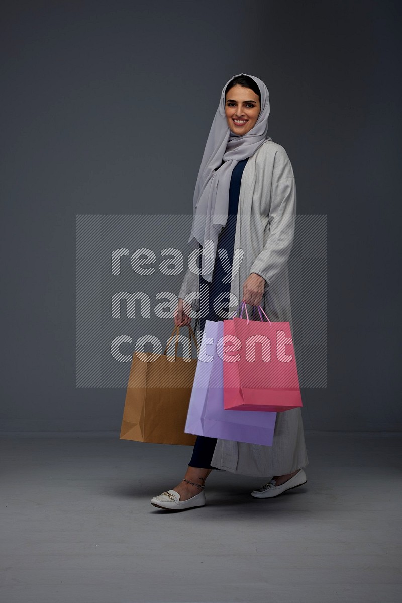 A Saudi woman wearing a light gray Abaya and head scarf standing and holding shopping bags on a grey background