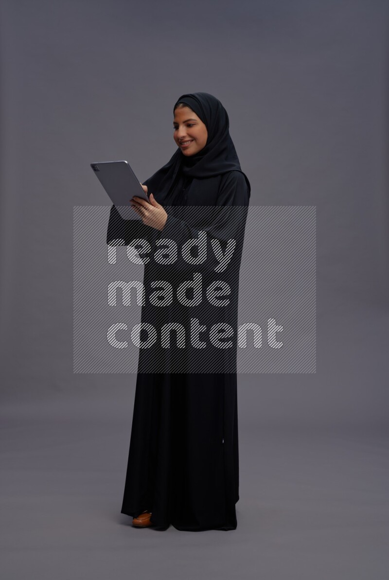 Saudi woman wearing Abaya standing working on tablet on gray background