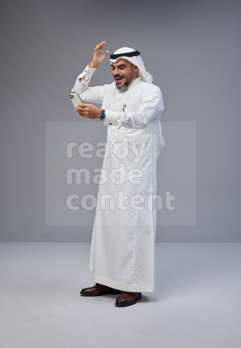 Saudi man Wearing Thob and white Shomag standing texting on phone on Gray background