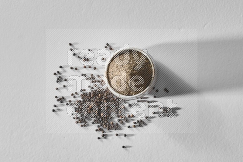 فلفل أسود مطحون في وعاء أبيض من السيراميك مع حبوب فلفل أسود علي خلفية بيضاء