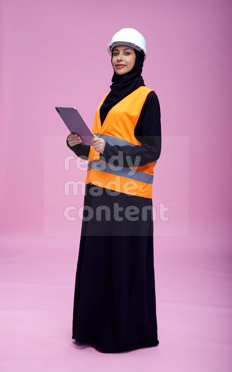 Saudi woman wearing Abaya with engineer vest and helmet standing working on tablet on pink background