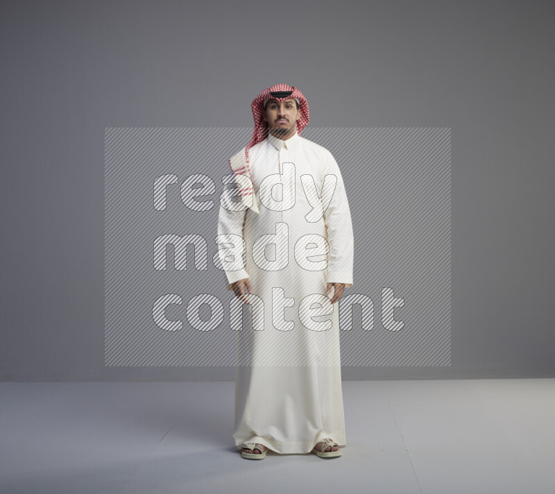 A Saudi man standing wearing thob and red shomag interacting with the camera on gray background