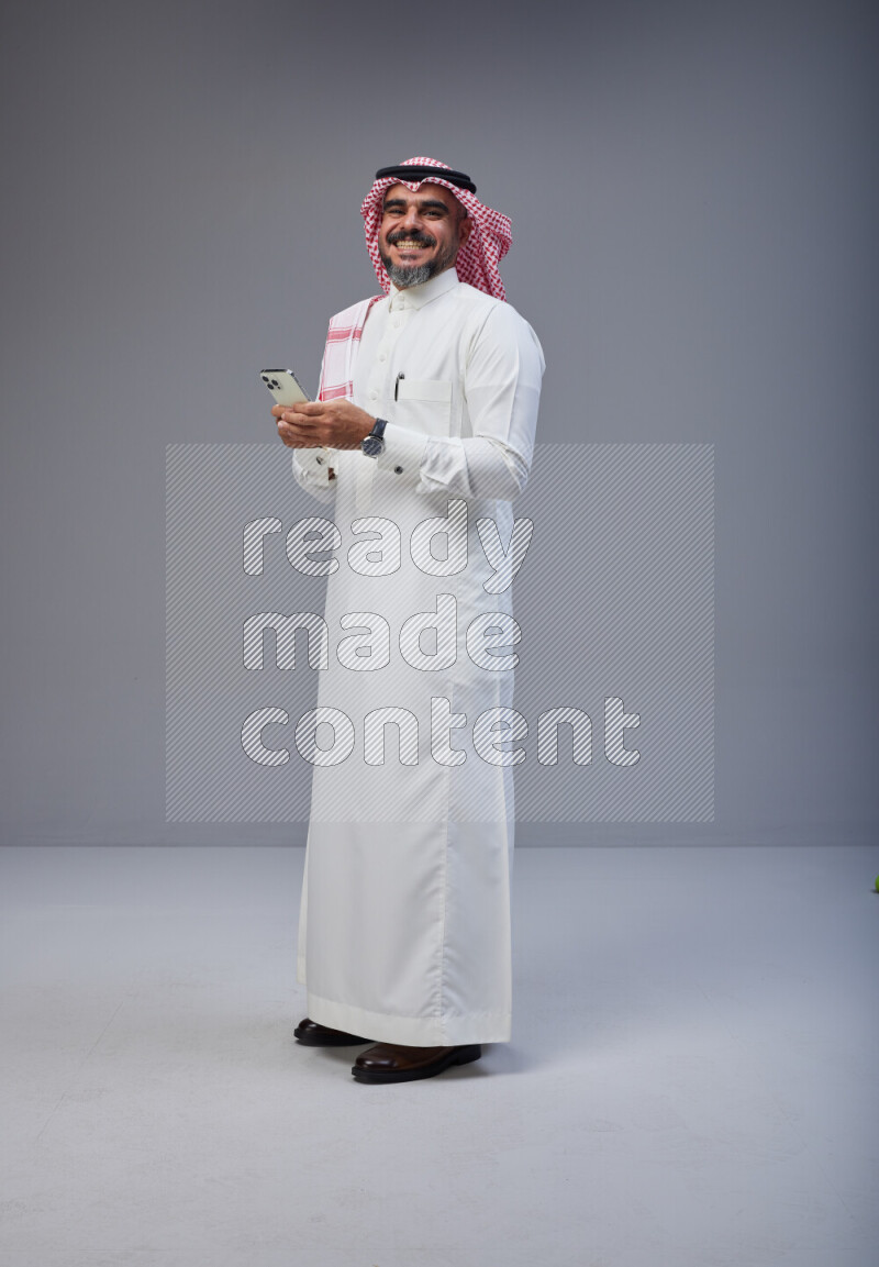 Saudi man Wearing Thob and red Shomag standing texting on phone on Gray background