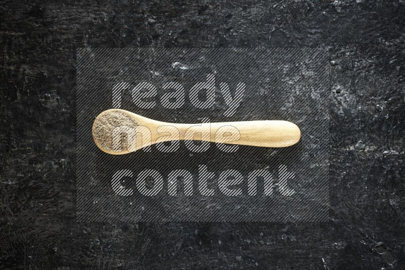 A wooden spoon full of cardamom powder on textured black flooring