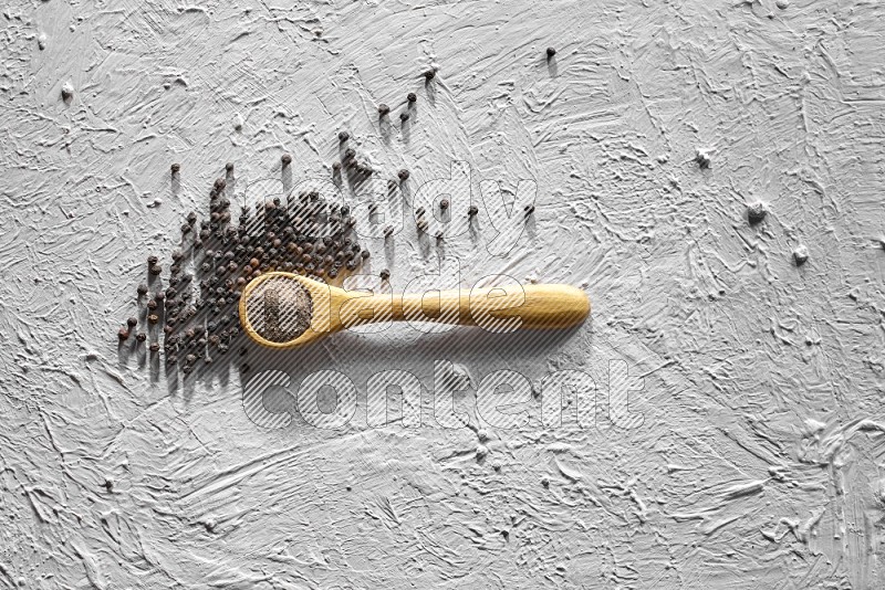A wooden spoon full of black pepper powder and black pepper beads beside it on a textured white flooring