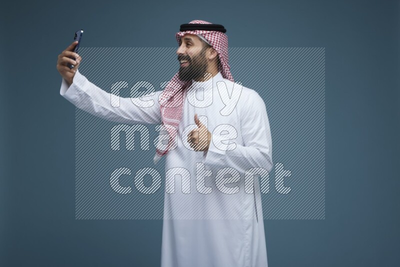 A man Taking Selfie in a blue background wearing Saudi Thob with Shomag