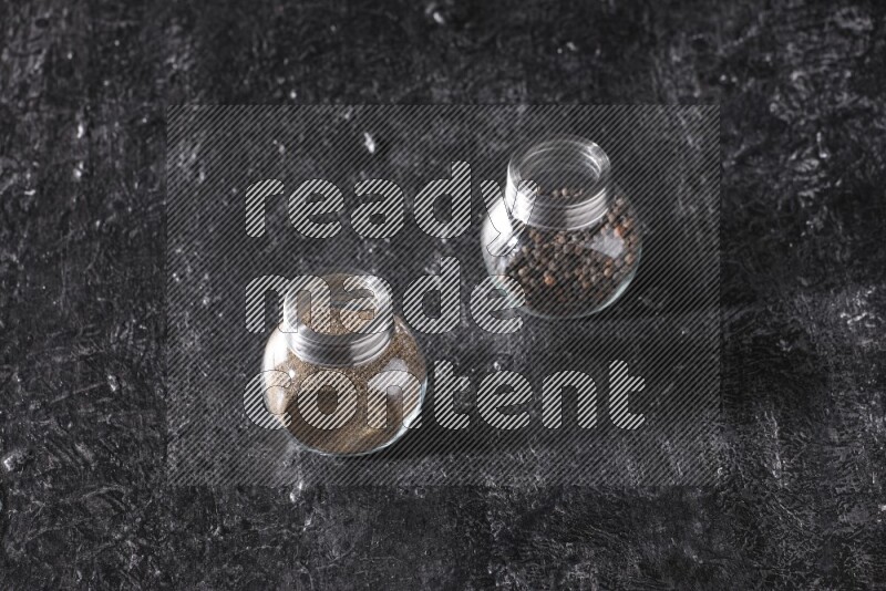 2 glass spice jars full of black pepper powder and black pepper beads on textured black flooring