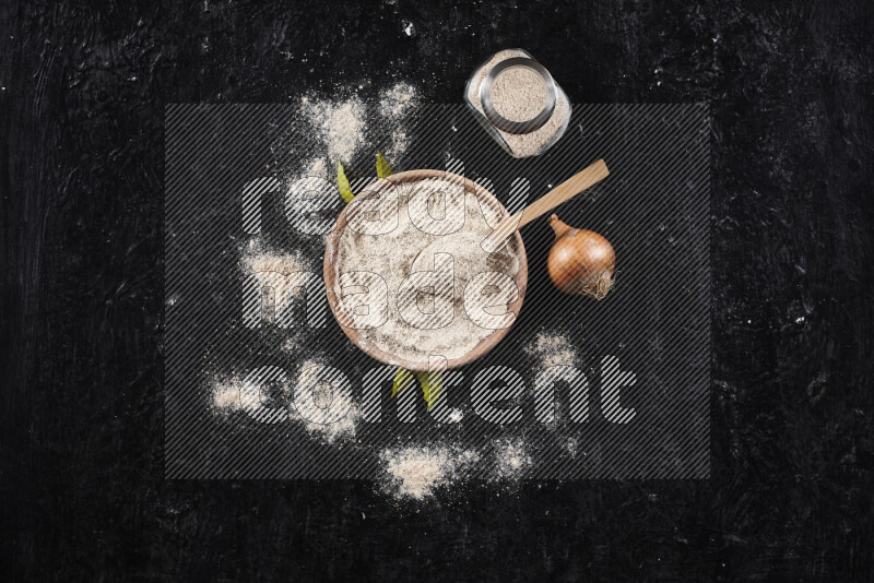 A wooden bowl full of onion powder with a glass jar beside it and fresh onion on black background