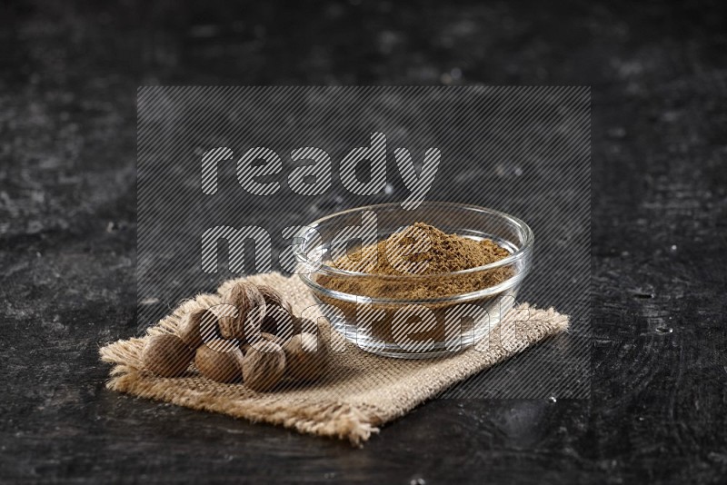 A glass bowl full of nutmeg powder with whole seeds beside it on burlap fabric on a textured black flooring